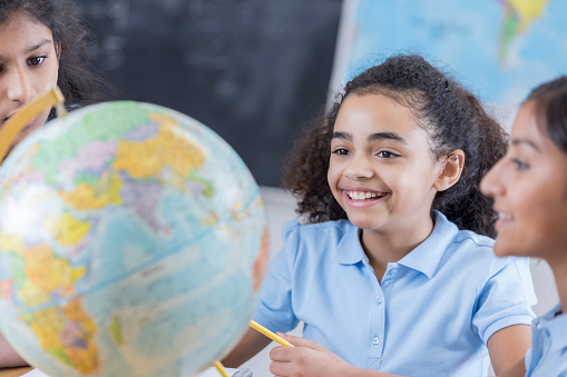 Smiling mixed race girl looks at a globe with her friends at school. They attend a private middle school.
