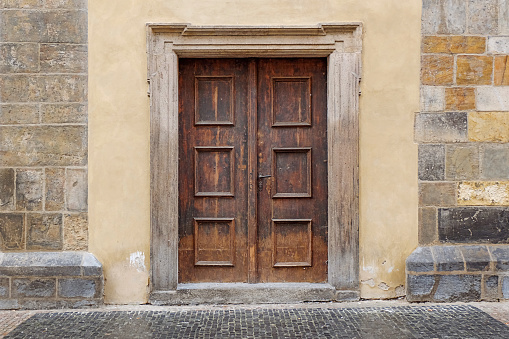 A brown wooden door entrance to an old building. The rectangle door is weathered and aged. The doorframe is made of stone with relief lines as ornaments. Around it is a yellow / beige facade and irregularly sized stone blocks.