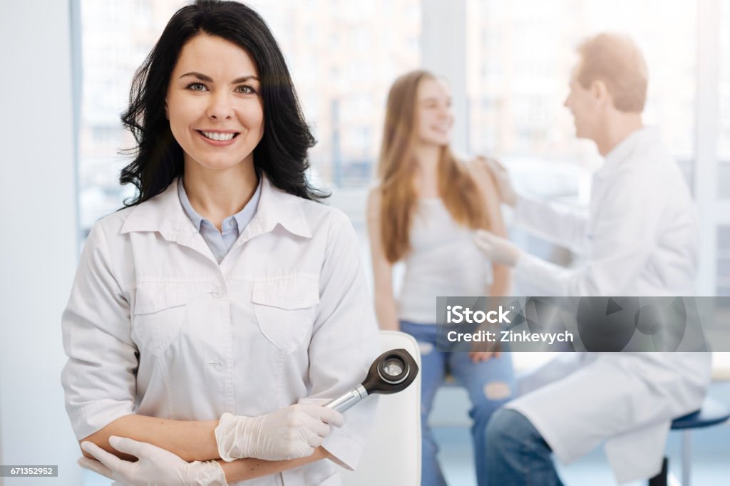 Happy young patient having conversation with dermatologist the hospital With individual attitude to every patient. Smiling charming beautiful dermatologist working in the clinic and holding dermatoscope while colleague talking to young patient in the background Dermatologist Stock Photo