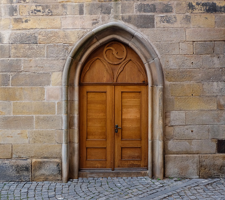 A brown wooden door entrance to an old building. The doorframe has a gothic style pointed arch. The wood is ornate with decorations above the entrance. It resembles being but is not part of an old castle or fortress.