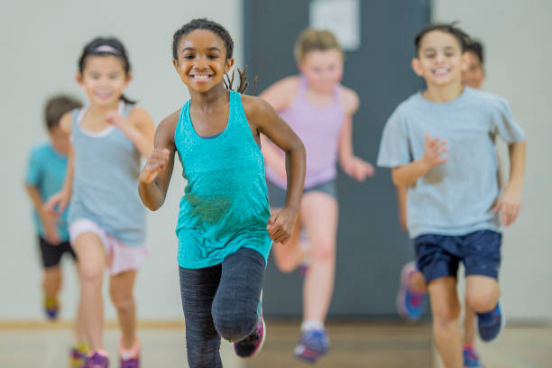 Jogging A group of elementary school children of various ethnicity are in PE class. They are indoors in the gym. They are playing a running game and smiling. A young girl of African descent is at the front. physical education stock pictures, royalty-free photos & images