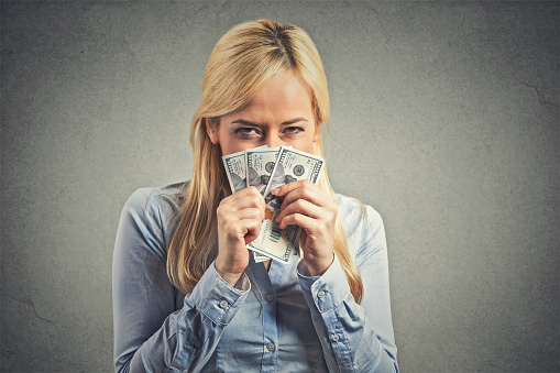 Closeup portrait greedy young corporate employee, woman, holding dollar banknotes tight isolated on gray wall background. Negative human emotion facial expression. Financial gain concept