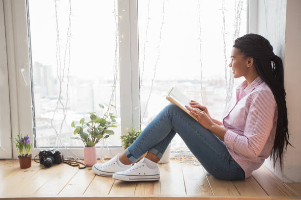 istock Dreamy female student with book sitting on window sill 671418220