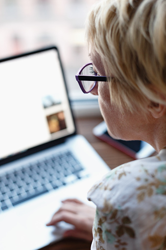 Back view of female in glasses sitting and working on laptop on blurred background.