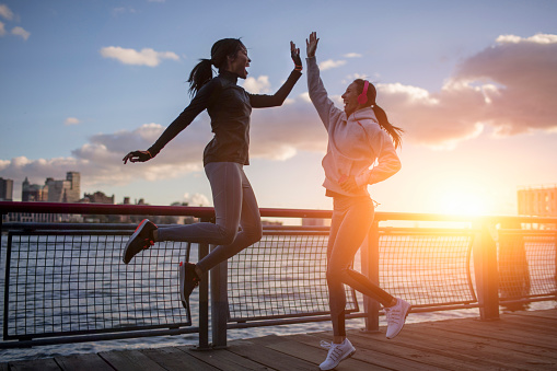 Full length of cheerful young women giving high-five during sunset. Multi-ethnic friends celebrating while jumping by East river. They are in sportswear.