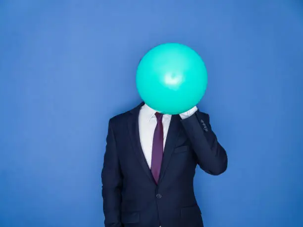 Young handsome bearded man in a suit blowing up a green balloon front view. Blue background.
