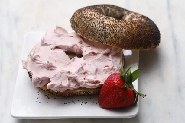 A high angle close up horizontal photograph of a sliced poppy seed bagel with strawberry flavored cream cheese and a fresh strawberry