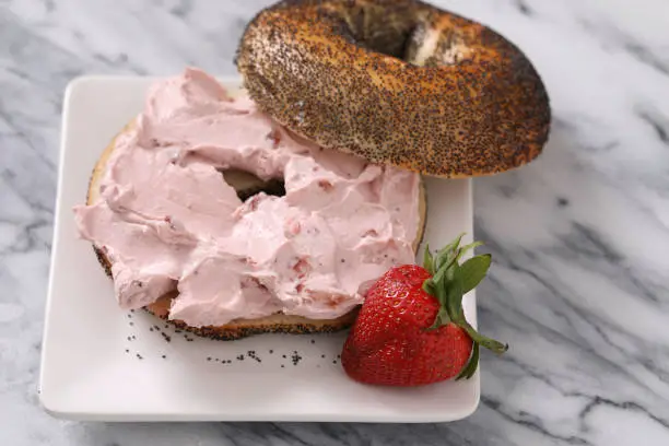 A high angle close up horizontal photograph of a sliced poppy seed bagel with strawberry flavored cream cheese and a fresh strawberry