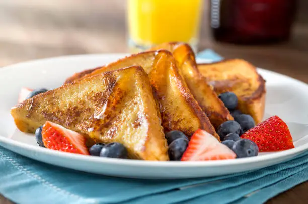 Plate of french toast in the morning light with maple syrup, blueberries, strawberries, and a glass of orange juice on rustic wood table with blue napkin.