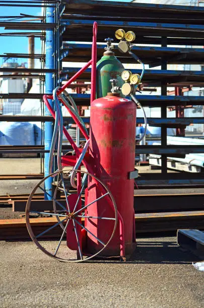 Acetylene torch for cutting steel and other metals. Tanks are sitting on old truck with hoses wrapped up in front of a variety of metal parts. In the background is a large factory. Outdoor setting, industrial setting.