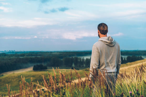 Man wears a grey hooded shirt and stands on field looking at the horizon. The sun is setting and the man stands with his back to the camera enjoying the view.