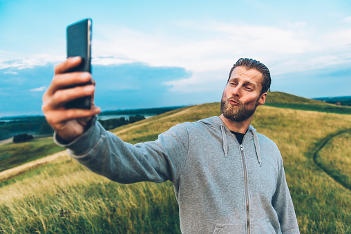 Man with beard wears a grey hooded shirt and stands while holding a 4G smart phone. He takes a selfie and makes a duck face look.