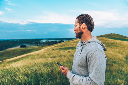 Man with beard wears a grey hooded shirt and stands on field looking at the horizon. He holds a 4G smart phone.