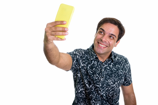Studio shot of young happy Persian man smiling while taking selfie picture with mobile phone isolated against white background horizontal shot