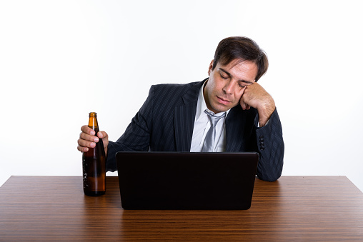 Studio shot of young Persian businessman looking drunk while holding bottle of beer and sitting with laptop on wooden table against white background horizontal shot