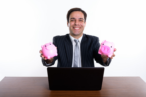 Studio shot of young happy Persian businessman smiling while holding two piggy banks with laptop on wooden table against white background horizontal shot