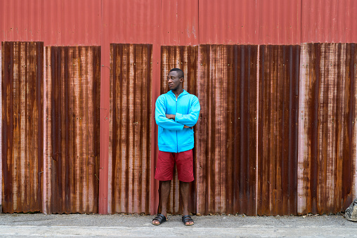Young black African man standing and thinking with arms crossed against old rusty sheet wall horizontal shot