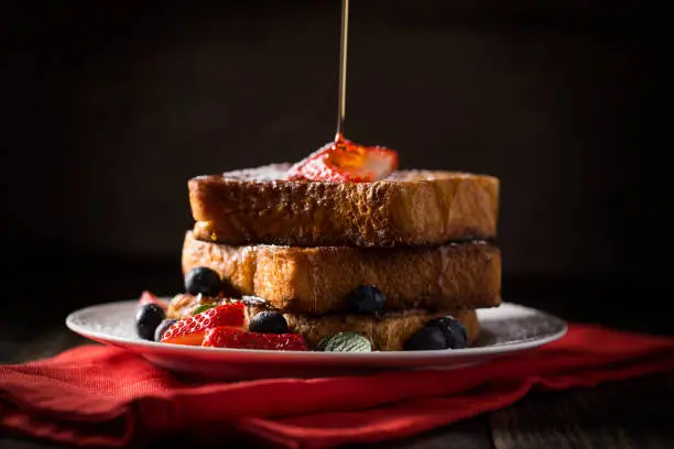 Maple syrup being poured over french toast with strawberries, blueberries, and mint on a rustic wood farm table.