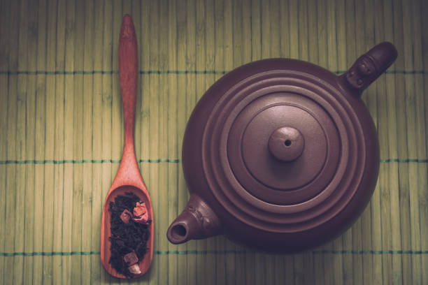 Close-up ceramic teapot, aromatic dry tea with fruits and petals in wooden spoon on  green bamboo background. Retro filter.