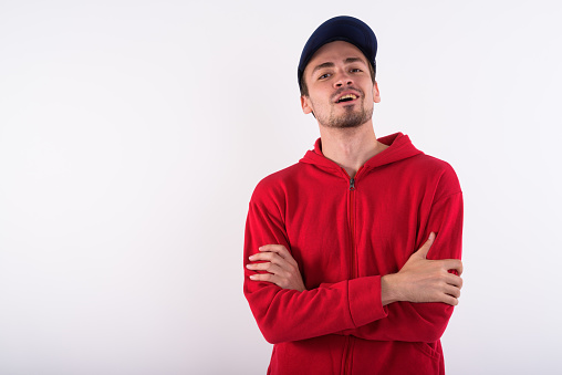 Studio shot of young happy bearded man smiling while wearing cap with arms crossed against white background horizontal shot