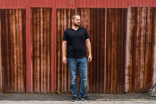 Young bearded man with hair tied standing and thinking against old rusty sheet wall horizontal shot