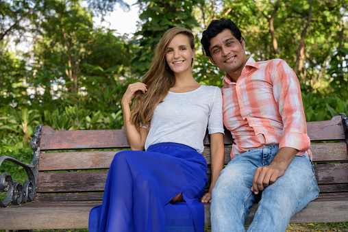 Happy multi ethnic couple smiling and sitting on wooden bench in love at peaceful green park horizontal shot