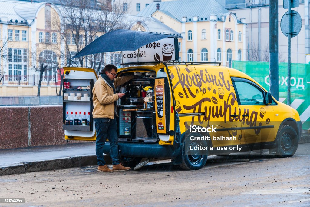 Mobile coffee bar in the rain MOSCOW, APRIL 16, 2017: Unidentified male vendor sales coffee and hot drinks on the street. Cold rainy day, no customers. The sign on the yellow car reads: Pushkin's coffee bar. Selling coffee on Moscow streets. Adult Stock Photo