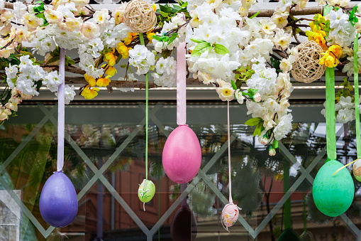 Colorful artificial easter eggs hang on ribbons in front of the glass window. Artificial flowers above eggs. Easter decoration. Deep blue, green, yellow, pink and red colors.