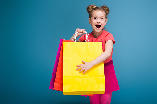 Isolated on blue, cute little caucasian brunette girl in pink dress hold purple, yellow and red paper bags, amazed, look at camera