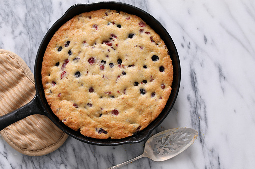 An overhead close up horizontal photograph of a freshly baked blueberry and cranberry skillet cake in a black cast iron skillet, a brown pot holder  and a metal serving spatula sit by the skillet.