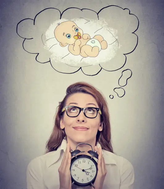 Photo of happy woman anticipating a baby looking up holding alarm clock