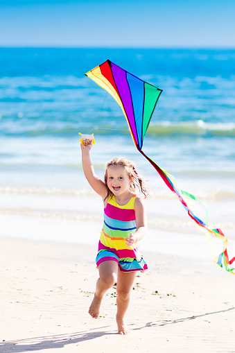 Happy laughing little girl flying a colorful kite running and jumping in sand on beautiful tropical beach during active summer family sea vacation. Kids play on ocean shore. Child with beach toys.