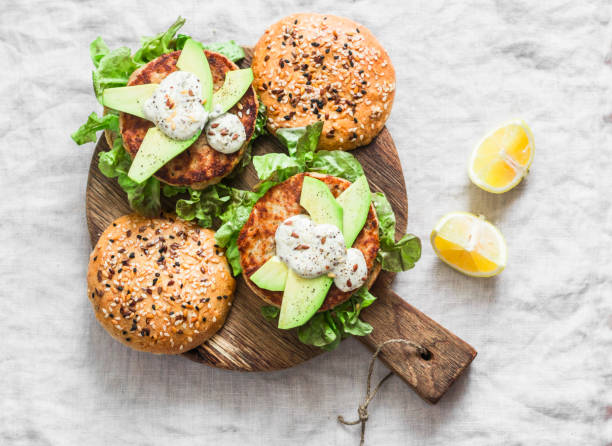 burger rybny. burgery z tuńczykiem, awokado i sosem musztardowym z pełnoziarnistymi domowymi bułeczkami na drewnianej desce do krojenia na lekkim tle, widok z góry - salad vegetable hamburger burger zdjęcia i obrazy z banku zdjęć