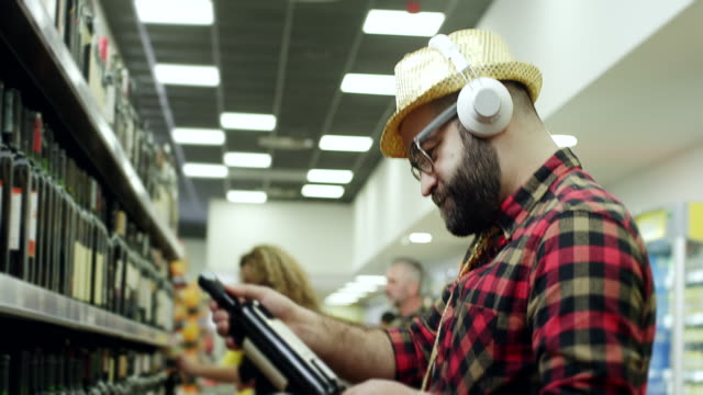 Man reading label on bottle of wine, listening music on headphones and vaping in supermarket