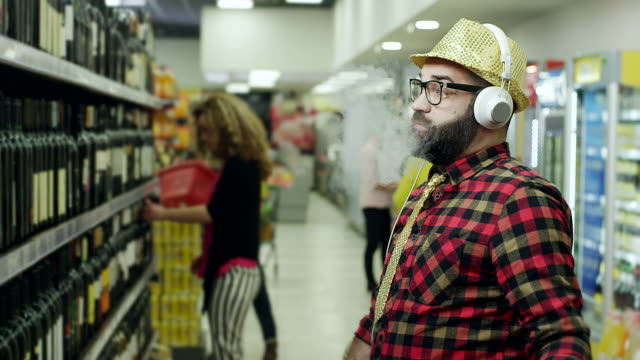 Men choosing bottle of wine, listening music on headphones and vaping in supermarket