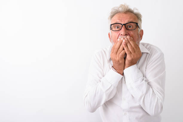 Studio shot of handsome senior bearded man looking shocked while covering mouth with both hands and wearing eyeglasses against white background