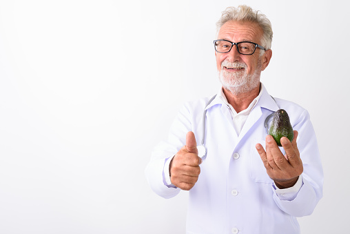 Studio shot of happy senior bearded man doctor smiling while holding avocado and giving thumb up against white background horizontal shot