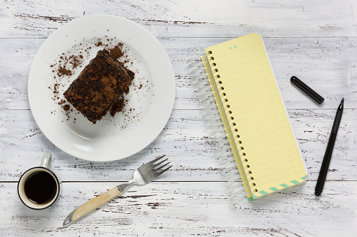 Chocolate cake, empty yellow notebook, coffee, fork and pen on wooden background
