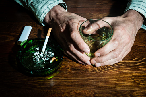 Hands of alcoholic man holding a glass with alcohol drink with smoking cigarette in the ashtray. Alcohol and cigarette addiction. Depression. Loneliness. Sadness.