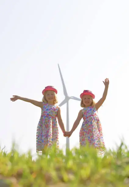 Twin sisters run carefree and happy towards the camera with a group of wind turbines behind them. Their faces express happiness for this type of environmental energy.