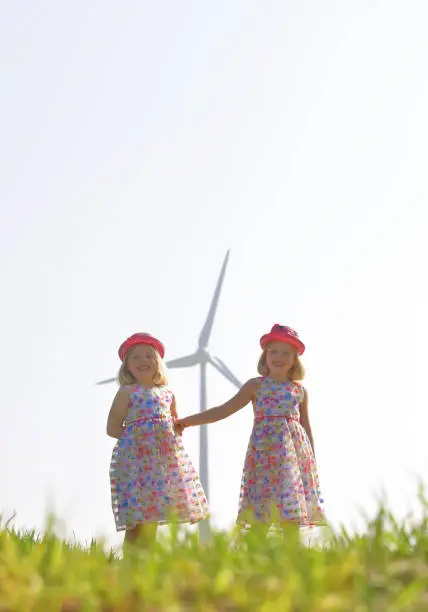 Twin sisters run carefree and happy towards the camera with a group of wind turbines behind them. Their faces express happiness for this type of environmental energy.