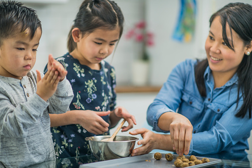An Asian mother, brother and sister are in a kitchen indoors. They are at the kitchen table and wearing casual clothing. They are baking chocolate chip cookies from scratch. They are rolling the cookies and putting them onto the baking tray.