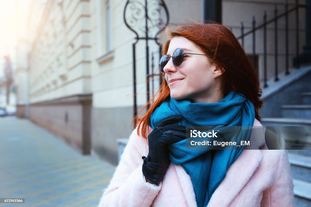 Garota linda de cabelo vermelha está andando em segundo plano urbano em um casaco cor de rosa e um lenço azul, com óculos de sol. - Foto de stock de Adulto royalty-free