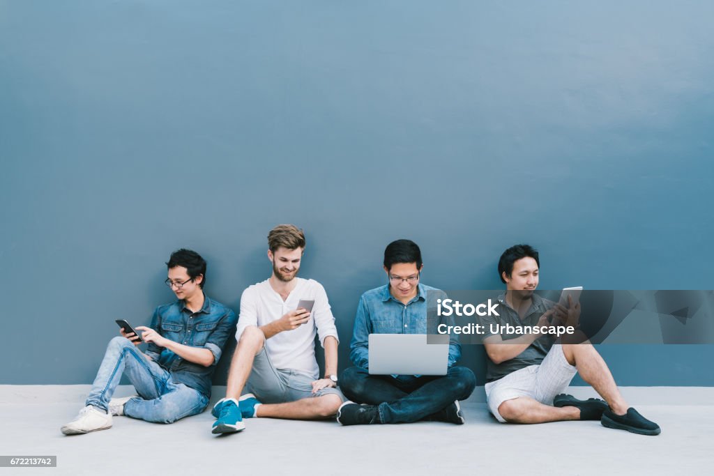 Multiethnic group of four men using smartphone, laptop computer, digital tablet together with copy space on blue wall. Lifestyle with infomation technology gadget, education, or social network concept Laptop Stock Photo