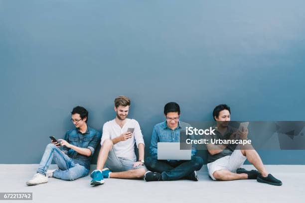 Multiethnic Group Of Four Men Using Smartphone Laptop Computer Digital Tablet Together With Copy Space On Blue Wall Lifestyle With Infomation Technology Gadget Education Or Social Network Concept Stock Photo - Download Image Now