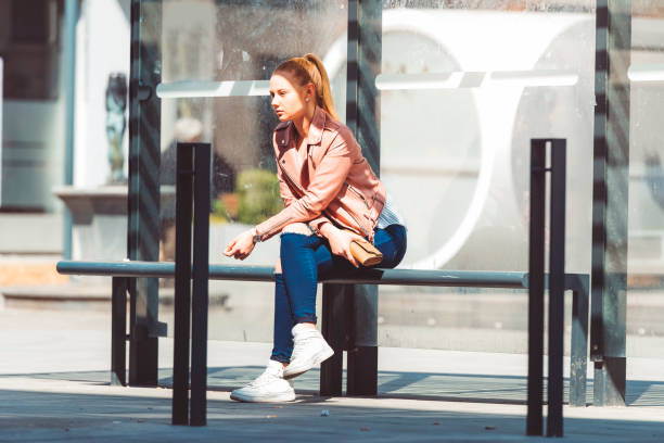 Young woman sitting and waiting at the bus station in the city centr on a sunny spring day