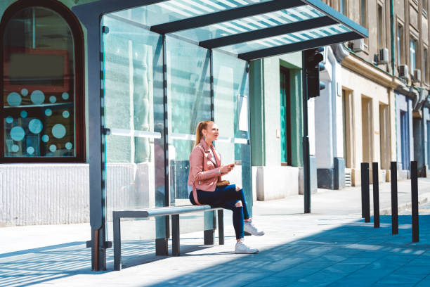 Beautiful young student at the glass made bus shelter Young woman sitting and waiting at the bus station in the city centr on a sunny spring day bus shelter stock pictures, royalty-free photos & images