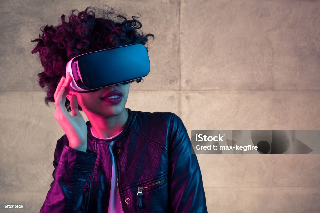 Young stylish girl in VR headset Young trendy female in leather jacket posing in VR glasses on background of concrete wall. Virtual Reality Simulator Stock Photo