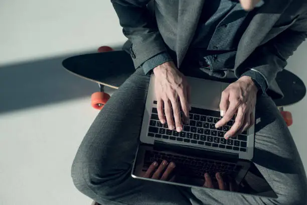 partial view of stylish businessman sitting on skateboard and typing on laptop