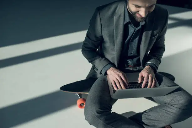 smiling stylish businessman sitting on skateboard and typing on laptop